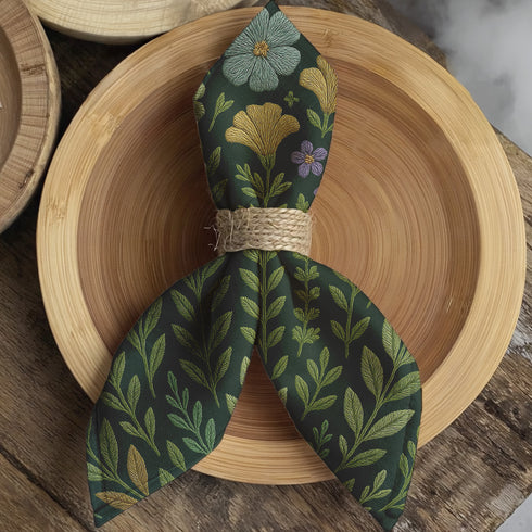 A 4-piece cloth napkin set featuring embroidered-style botanical illustrations of wildflowers and herbs in shades of gold, mint, and lavender on a dark green background; made from soft polyester, square-shaped, with a luxe and mystical design.